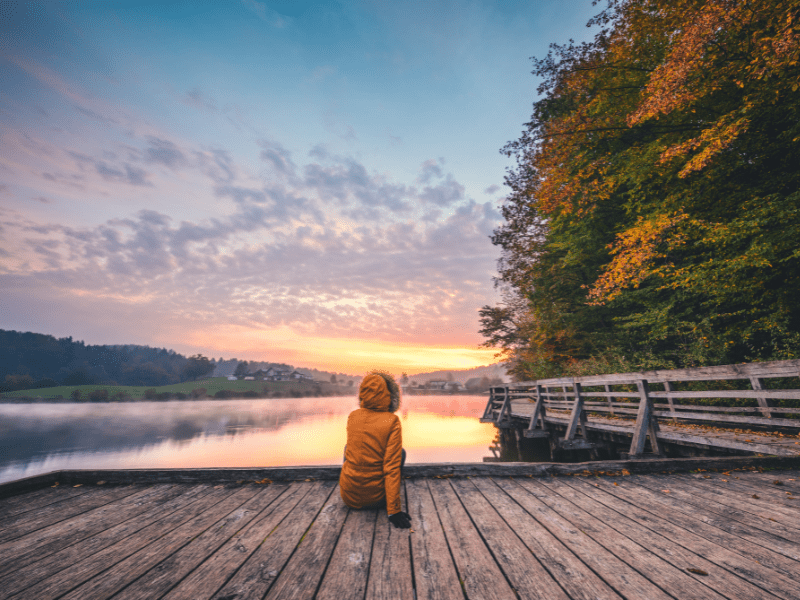 A person sitting on a wooden dock. Looking at the sunset sky behind the image.